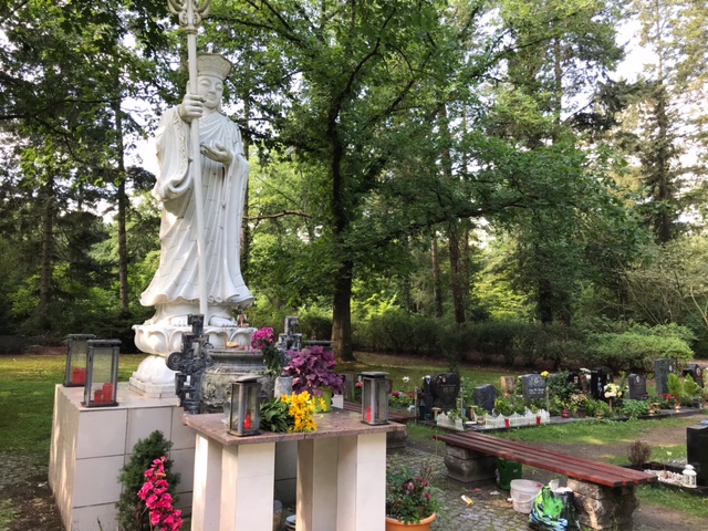 A tall white Buddha statue in a quiet cemetery with many trees. The statue is surrounded by red candles.