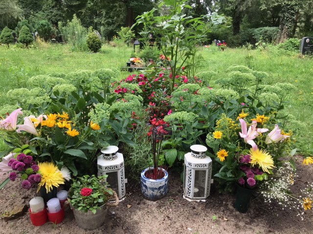 In der Mitte befinden sich Räucherstäbchen, Kerzen und Kerzenhalter, umringt von Blumensträußen. Im Hintergrund ist der grüne Friedhof.