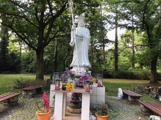 Vorderansicht der weißen Buddha Statue. Auf dem Podest der Statue stehen Blumen und Kerzenhalter. Rechts und links sind Holzbänke. Im Hintergrund ist der Wald.