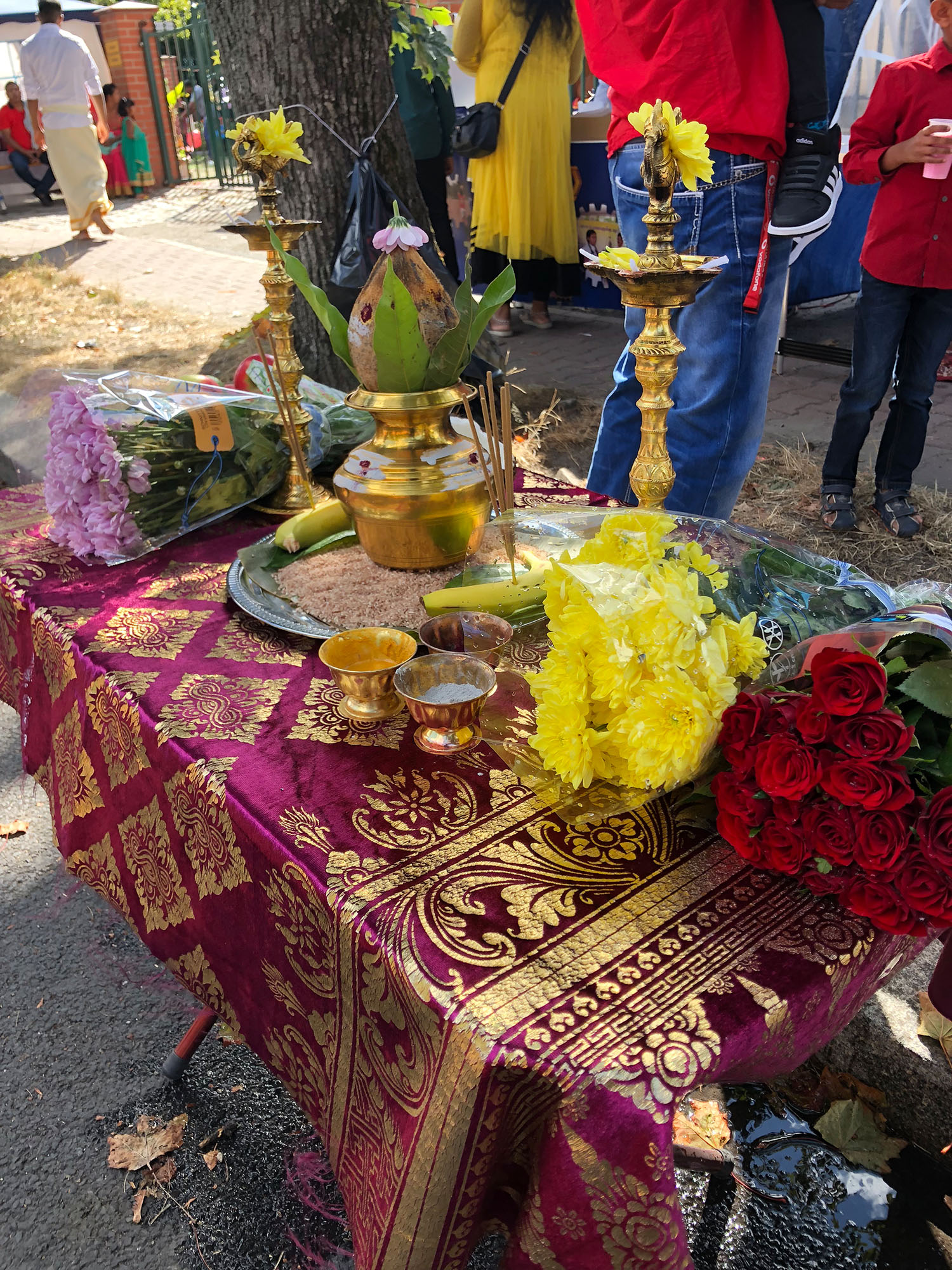 A table with a purple and gold decorated tablecloth. On the table are tall lamps filled with oil on either side, which are lit before making special offerings to God. In the center is a vessel filled with water that is sprinkled in homes after special services as a sign of holiness. The top of the vessel is covered with coconut and mango leaves.