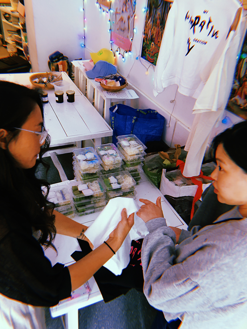 Vicky on the left and another person on the right are looking at T-shirts. They are standing in the store and in the background you can see packaged food and more T-shirts.