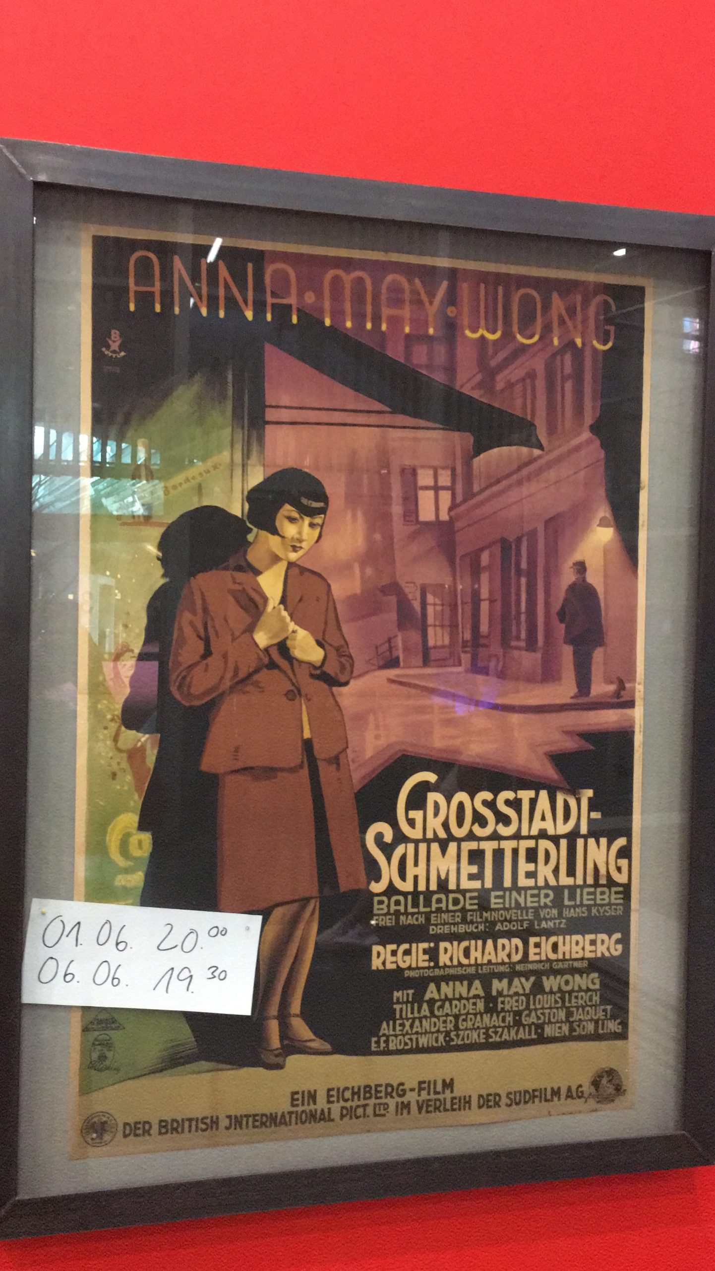 A framed, illustrated movie poster with Anna May Wong in the foreground. In the background one can see an alley and the outlines of another person. At the top of the poster it says “Anna May Wong”. Below is the caption “Großstadtschmetterling” (translated: Big City Butterfly). On the frame of the movie poster is a white note with black writing, it says “01.06. 8pm, 06.06. 7.30 pm”.