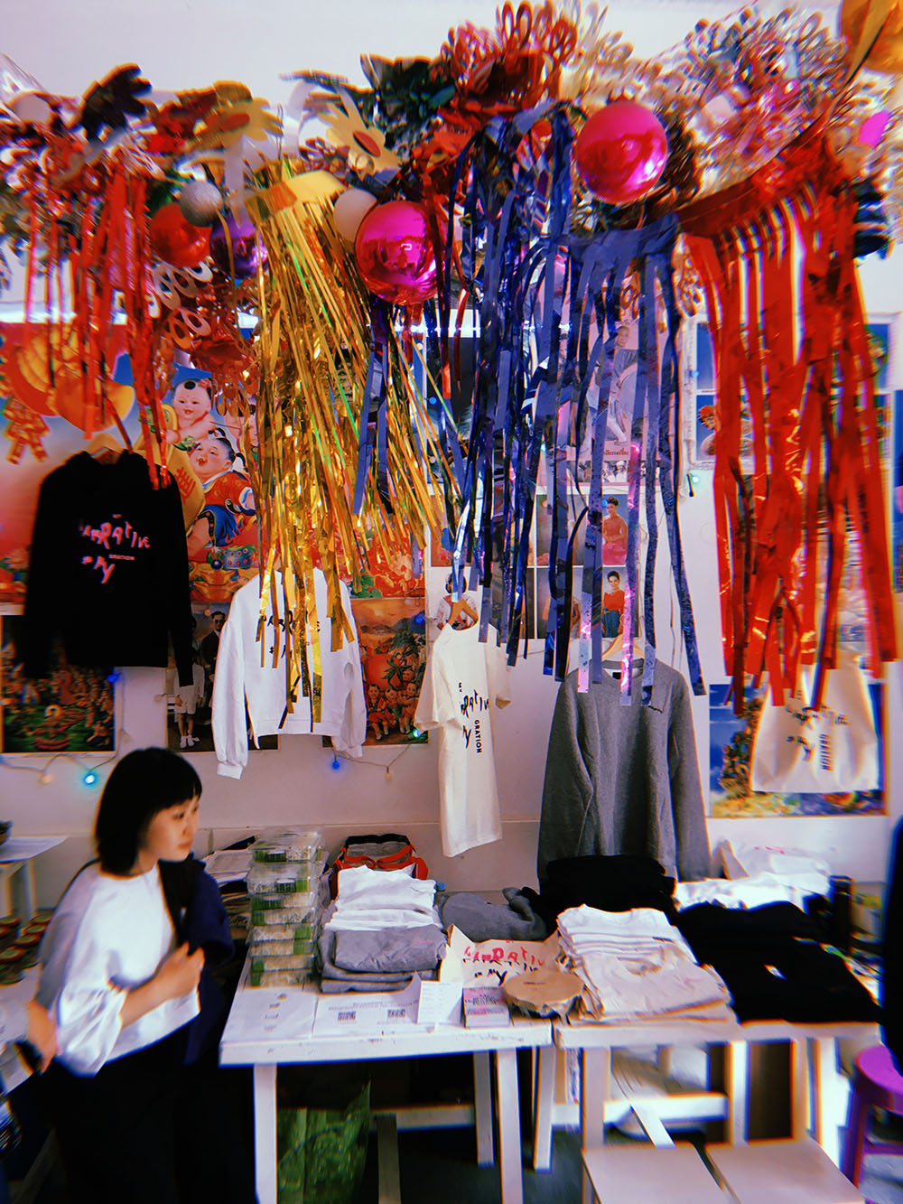 The Mygration festival is holding an event in the Pandanoodle store on the 20.06.2020. There are colorful, glittery garlands hanging above and T-shirts for sale on the wall. A person sits at the table to the left. 