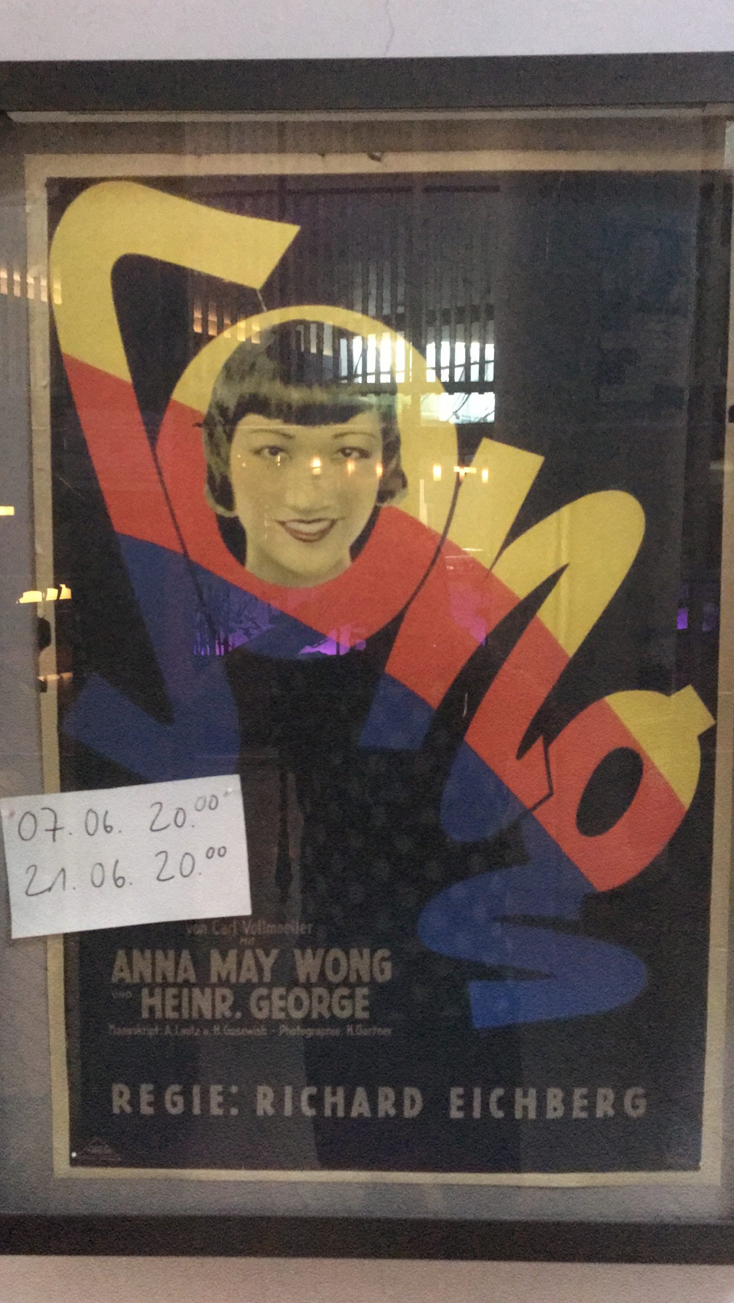 A framed, black movie poster with yellow-red-blue headline across the whole poster: “SONG”. Anna May Wong's face fills the “O” in “SONG” from inside. At the bottom it reads “Anna May Wong, Heinr. George. Regie Richard Eichberg” (translated: Anna May Wong, Heinr. George. Directed by Richard Eichberg). To the left above the frame is a white slip of paper with black writing, it says “07.06. 8pm, 21.06. 8pm”.