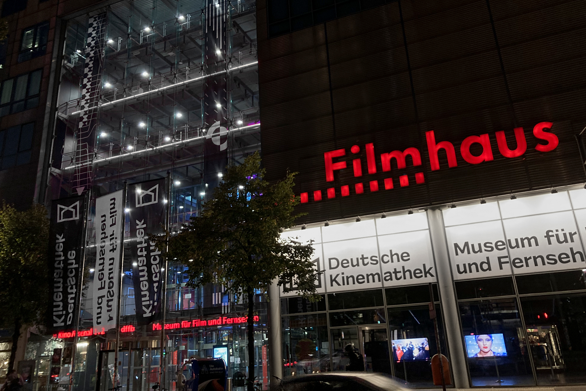 Front view from the Filmhaus in Berlin with the red lit-up label “Filmhaus”. Below are two white lit-up boards with black color “Deutsche Kinemathek” (left side) and “Museum für / und Fernseh” (right side, cut).