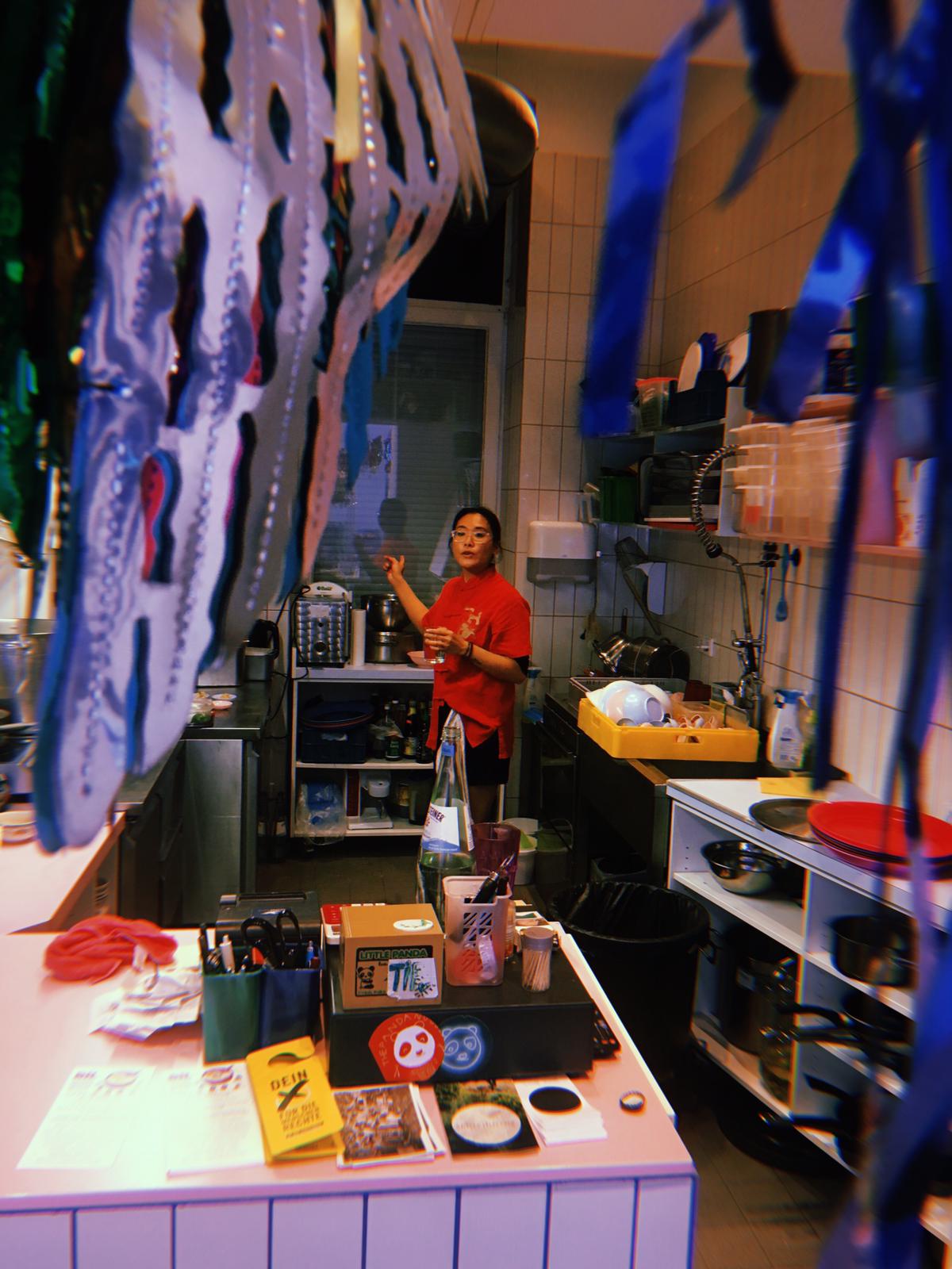 Vicky trägt ein rotes T-Shirt und steht in der Küche von dem Pandanoodle Laden. Im Vordergrund hängen bunte Girlanden. Das Foto wurde während des Rice is Life Events am 19.05.2019 aufgenommen.