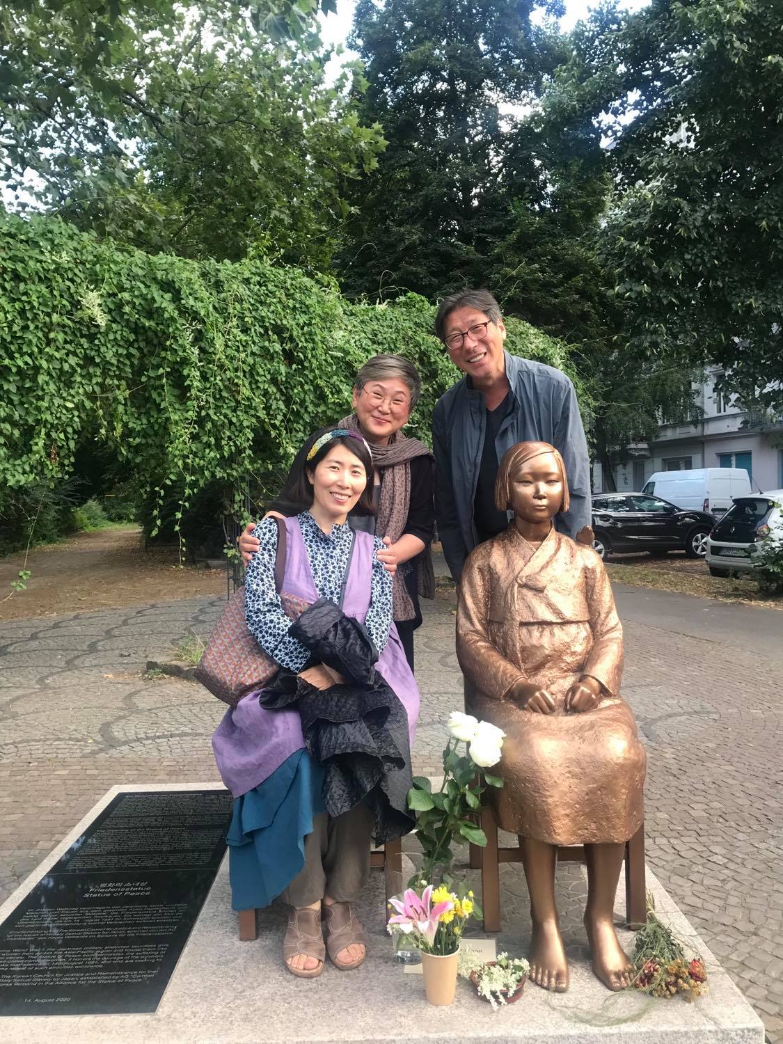Die Friedensstatue in gold hat die Gestalt eines jungen Mädchens. Links neben ihr sitzt eine Frau. Hinter den beiden stehen zwei weitere Menschen. Im Hintergrund fängt der Park an.