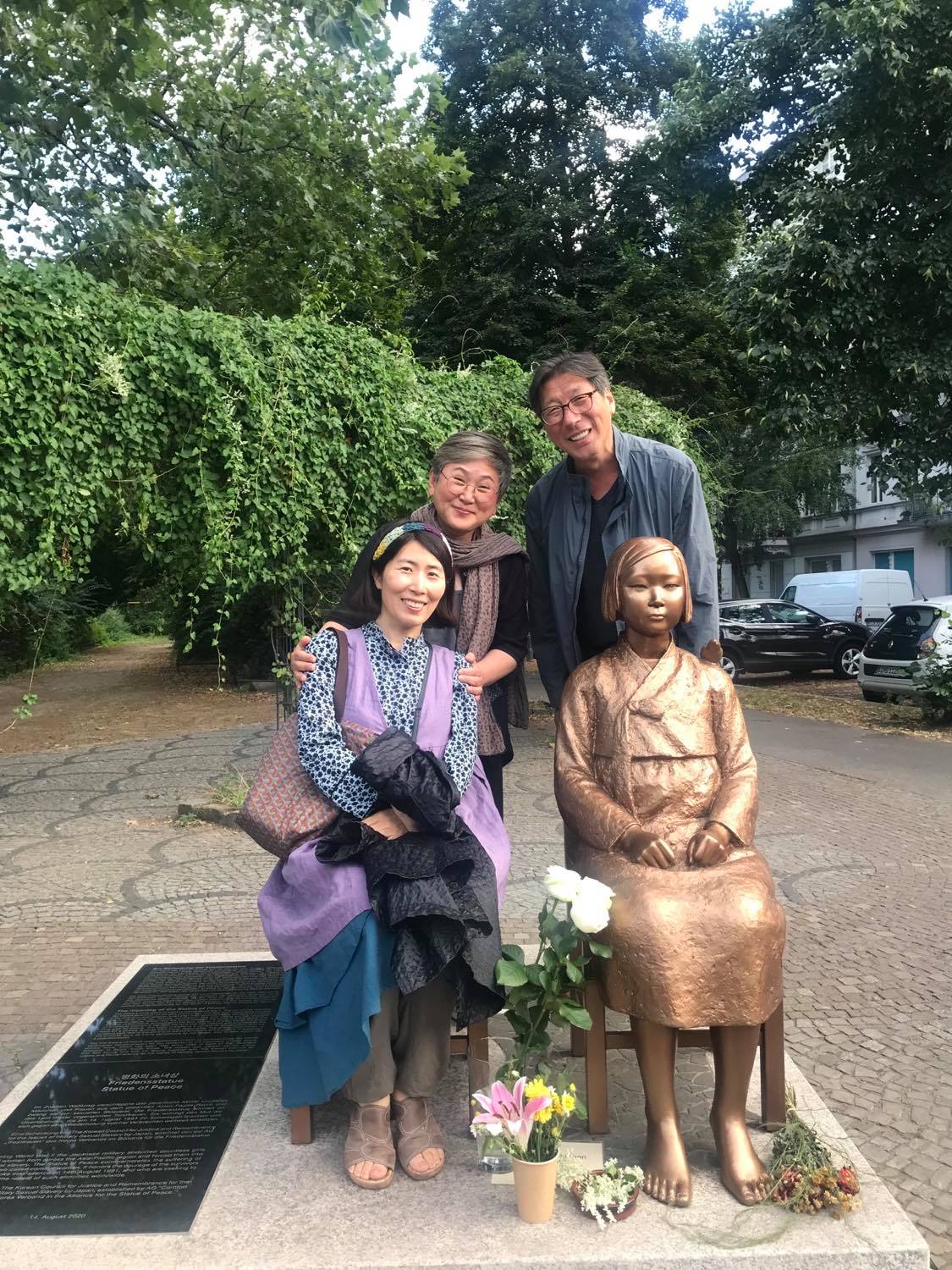 Statue of peace in gold has an appearance of a young girl sitting on a chair. To her left is a woman. Behind them are two other people. In the background is the park.