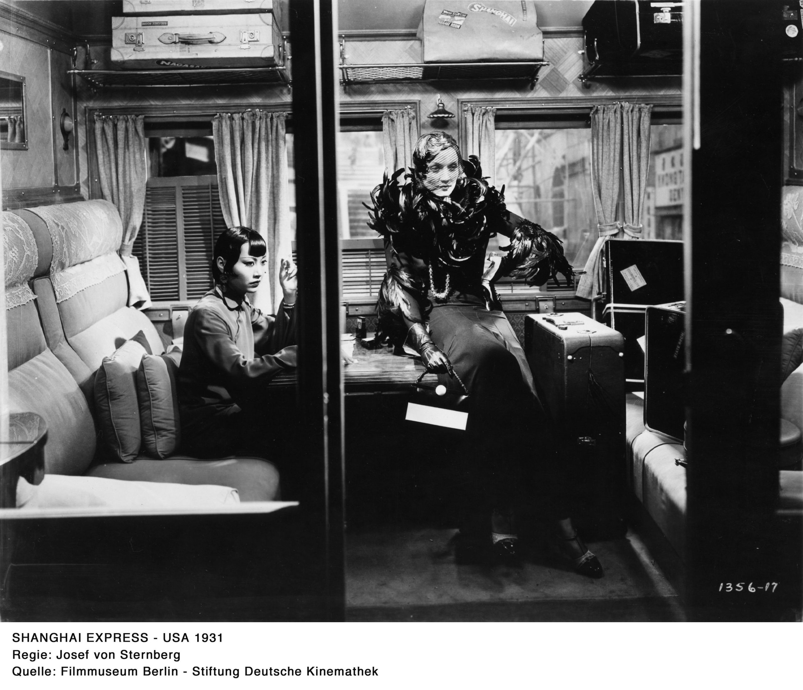 Ein schwarz-weiß Filmausschnitt aus dem Film “Shanghai Express”. Anna May Wong sitzt links auf einem Sitzplatz in einem Zugabteil. Eine weiße Frau in eleganter Kleidung lehnt an einem Tisch, rechts daneben ist ein großer Koffer.